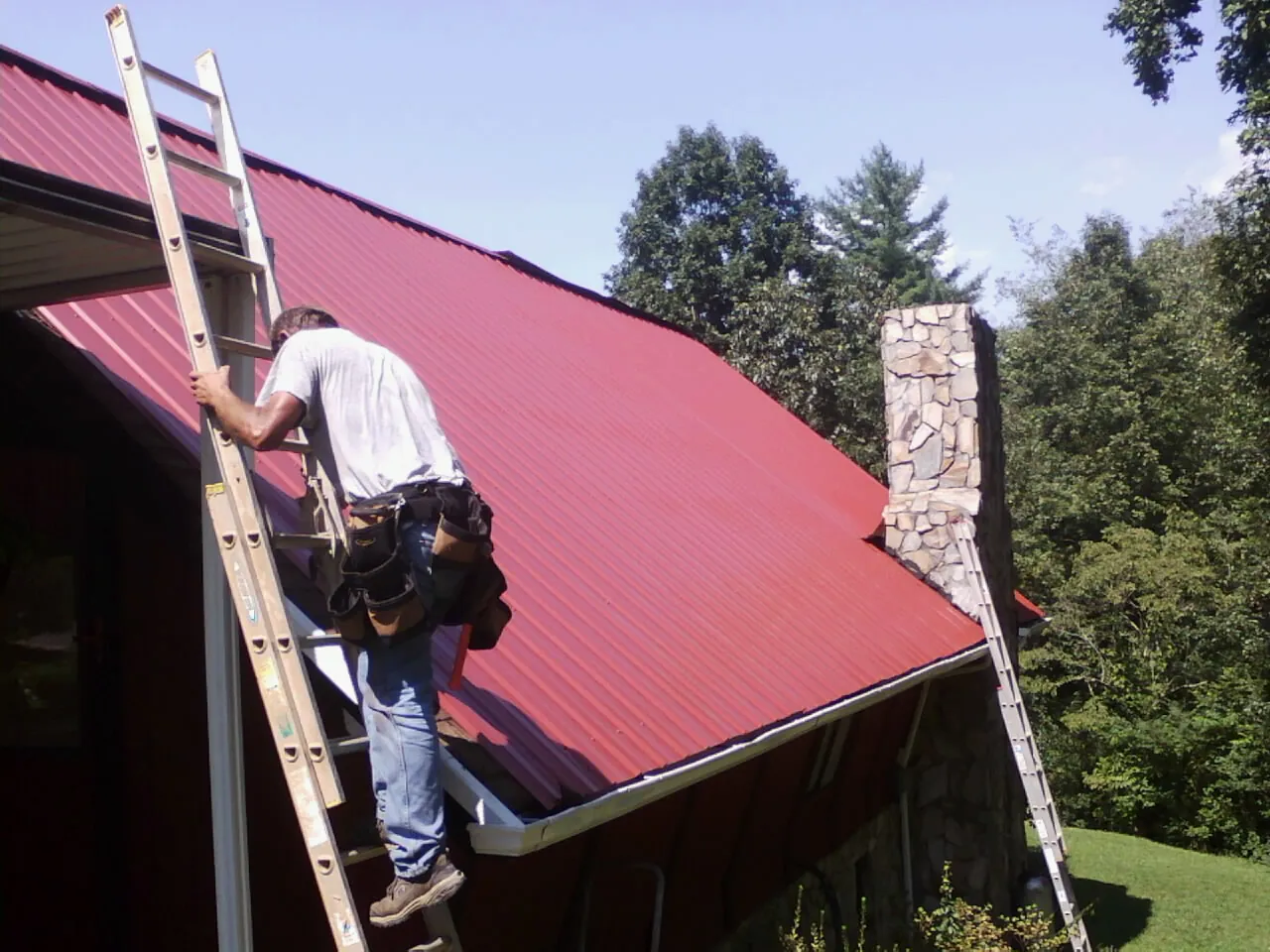 Roofer on a ladder beside a red metal roof and stone chimney — Soffit & Fascia Repair by Roofing Brothers in Milano